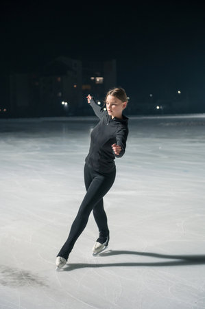 A young woman in black athletic wear gracefully ice skates on a lit outdoor rink at night, surrounded by soft lighting.の写真素材