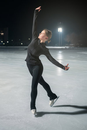 A young woman in black athletic wear gracefully ice skates on a lit outdoor rink at night, surrounded by soft lighting.の写真素材