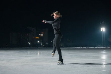 A young woman in black athletic wear gracefully ice skates on a lit outdoor rink at night, surrounded by soft lighting.の写真素材