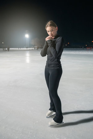 A young woman in black athletic wear gracefully ice skates on a lit outdoor rink at night, surrounded by soft lighting.の写真素材