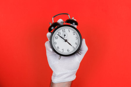 A white-gloved hand holds a classic black alarm clock against a bold red background, highlighting time management and punctuality.の写真素材