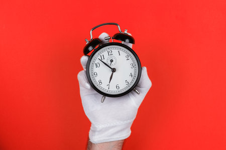 A white-gloved hand holds a classic black alarm clock against a bold red background, highlighting time management and punctuality.の写真素材