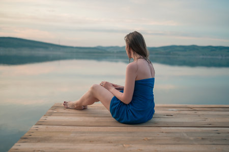 A woman in a blue towel sits on a wooden dock, gazing at the calm lake and peaceful natural surroundings.の写真素材