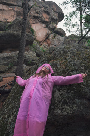 A woman wearing a pink raincoat leans against a mossy rock, embracing the wilderness of a lush, rocky forest setting.の写真素材