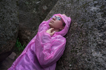 A woman wearing a pink raincoat leans against a mossy rock, embracing the wilderness of a lush, rocky forest setting.の写真素材