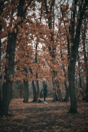 A woman dressed in a beanie and jacket standing in a forest with golden autumn leaves, conveying a cozy seasonal vibe.の写真素材