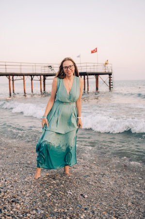A woman in a flowing green dress poses on a pier by the sea at sunset, with a Turkey flag waving in the background.の写真素材