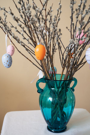 Decorative Easter eggs in various pastel colors hang on pussy willow branches arranged in a vase,...