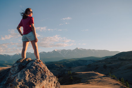A woman in casual clothing stands on a rocky peak, gazing at scenic mountains and a vast valley under golden sunlight.の写真素材