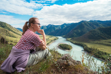 A woman in casual outdoor clothing sits on a hillside, admiring a breathtaking view of mountains, a winding Katun river, and lush greenery. Trip on Altai Mountains in Altai Republicの写真素材