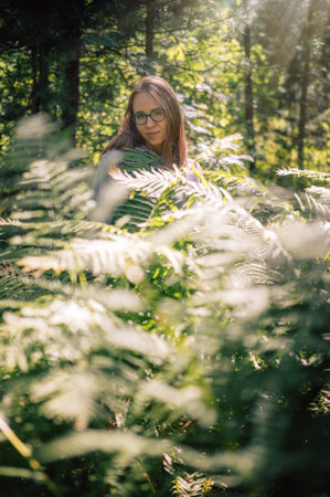A thoughtful woman in glasses stands amidst lush green ferns in a sunlit forest, enjoying the tranquility of nature.の写真素材