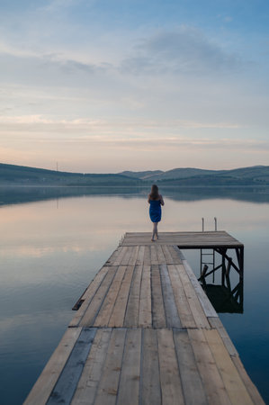 A young woman with wet hair, wrapped in a blue towel, sits on a wooden dock by a calm lake during sunset.の写真素材