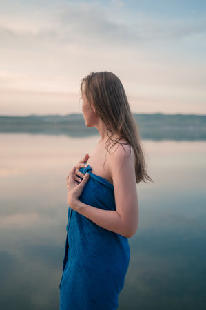 A young woman with wet hair wrapped in a blue towel stands near a calm lake at sunset, gazing thoughtfully into the distance.の写真素材