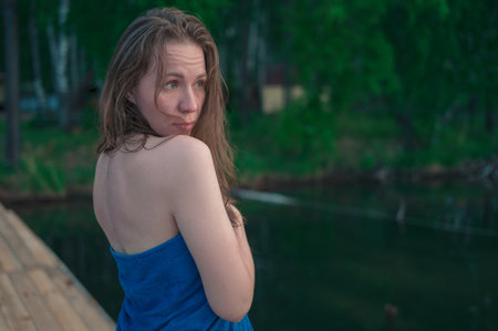 A woman with wet hair wrapped in a blue towel stands near a calm lake at sunset, gazing thoughtfully into the distance.の写真素材