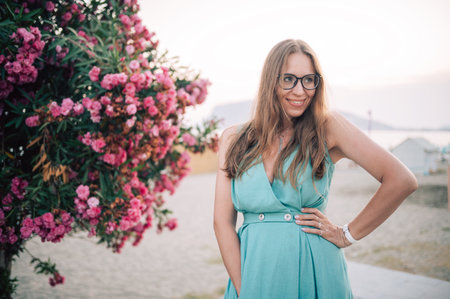 A graceful woman in a turquoise dress poses near blooming pink flowers, enjoying a peaceful moment in a natural outdoor setting.の写真素材