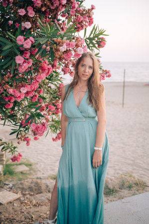 A graceful woman in a turquoise dress poses near blooming pink flowers, enjoying a peaceful moment in a natural outdoor setting.の写真素材