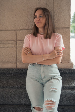 Young woman in a pink t-shirt and ripped jeans leans outdoor, looking thoughtful.の写真素材