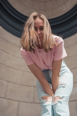 Young woman in a pink t-shirt and ripped jeans leans outdoor, looking thoughtful.の写真素材