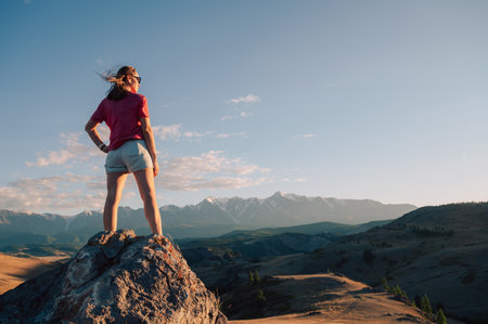 A woman in casual clothing stands on a rocky peak, gazing at scenic mountains and a vast valley under golden sunlight.の写真素材