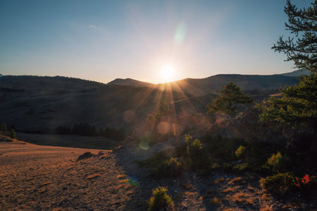 A breathtaking sunset over a rugged mountain landscape, casting warm golden light across hills, trees, and rocky terrain. Altai Mountainsの写真素材