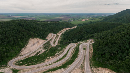 Aerial view of a curvy road in Belokurikha, Altai, cutting through lush green mountains and forested slopes, surrounded by farmland and countryside.の写真素材