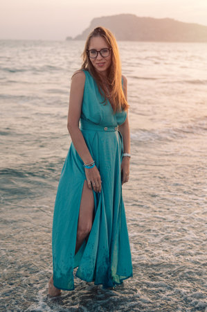 A woman in a flowing green dress poses on a pier by the sea at sunset.の写真素材