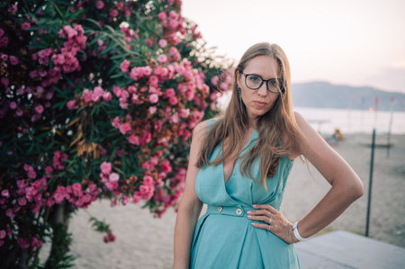 A graceful woman in a turquoise dress poses near blooming pink flowers, enjoying a peaceful moment in a natural outdoor setting.の写真素材