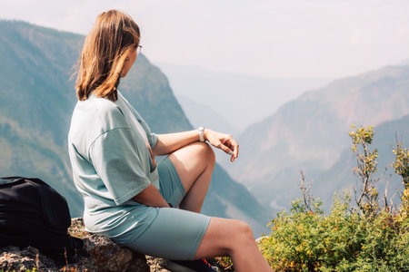 Young woman in sportswear sits on a rock ledge, peacefully observing scenic mountain landscape during a summer hiking adventure.の写真素材