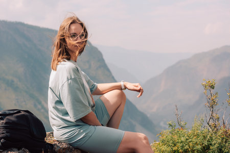 Young woman in sportswear sits on a rock ledge, peacefully observing scenic mountain landscape during a summer hiking adventure.の写真素材