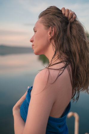 A young woman with wet hair wrapped in a blue towel stands near a calm lake at sunset, gazing thoughtfully into the distance.の写真素材
