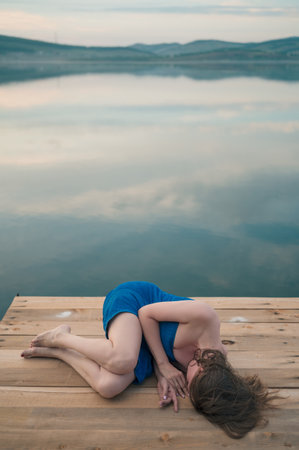 A woman with wet hair, wrapped in a blue towel, lie on a wooden dock by a calm lake during sunset.の写真素材