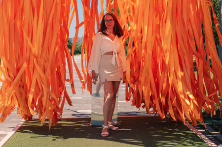 Stylish woman in a white outfit standing confidently in an outdoor installation with long, flowing orange ribbons in sunlight.の写真素材
