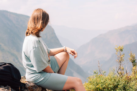 Young woman in sportswear sits on a rock ledge, peacefully observing scenic mountain landscape during a summer hiking adventure.の写真素材