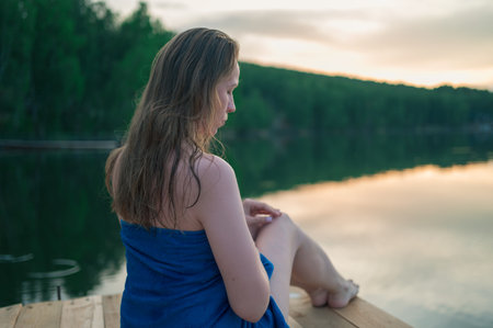 Young woman with wet hair sitting on a wooden pier wrapped in a blue towel, enjoying peaceful lake scenery at sunset.の写真素材