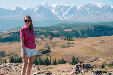 A woman in casual clothing stands on a rocky peak, gazing at scenic mountains and a vast valley under golden sunlight.の写真素材