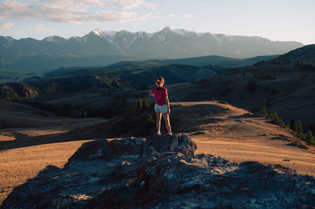 A woman in casual clothing stands on a rocky peak, gazing at scenic mountains and a vast valley under golden sunlight.の写真素材