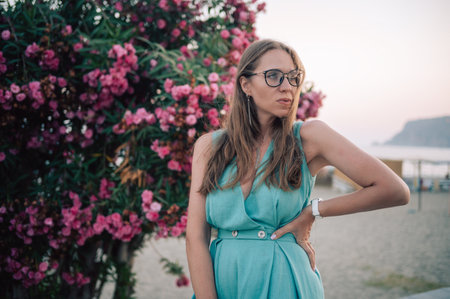 A graceful woman in a turquoise dress poses near blooming pink flowers, enjoying a peaceful moment in a natural outdoor setting.の写真素材