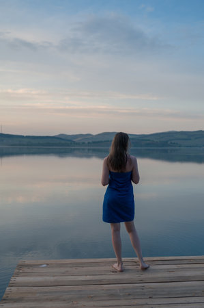 A woman on a wooden dock by a calm lake during sunset.の写真素材