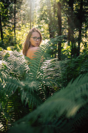 Back view of a woman standing in a lush green forest, touching her hair while sunlight filters through the trees and ferns.の写真素材