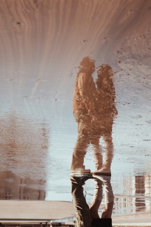 Reflection in puddle of woman and man embracing under umbrella during rainの写真素材
