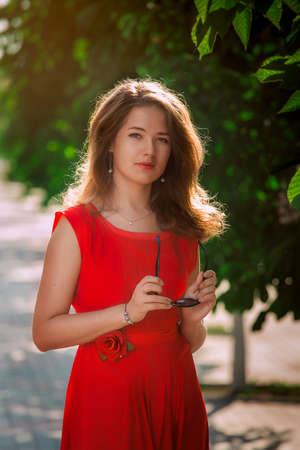 Portrait of a beautiful sexy fashion girl in a red dress on the nature of the park, with dark hair and a leather beltの写真素材