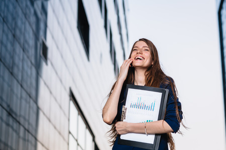 Successful smiling businessman, standing against the backdrop of buildings  holding  folder with sales charts. City business woman working.の写真素材