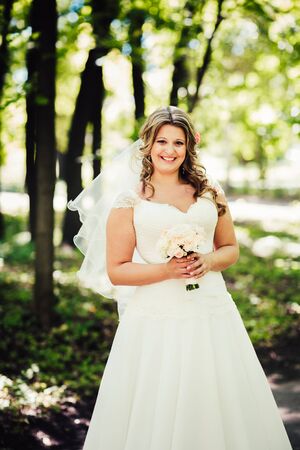 Stylish beautiful happy bride holding   bouquet of flowers on a background of trees in the summer park close-upの写真素材