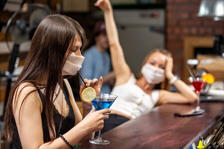 girls in medical masks chatting in a pub. girls in medical masks are talking at the bar. girls in medical masks drink cocktailsの写真素材