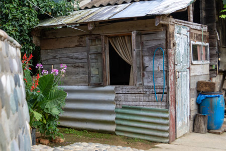 Old wooden house with corrugated metal panels, open window and door, flowers and a blue barrel in a rural yard, showing simple living and rustic village life