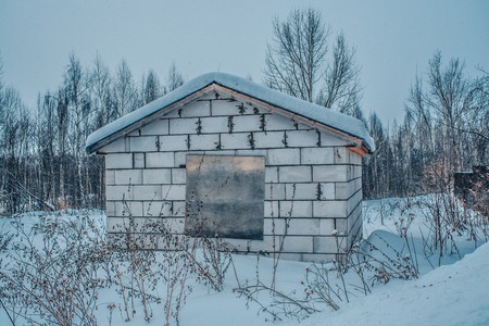 Old barn grey concrete wall with snow on the roof and boarded up the window on a forest backgroundの写真素材