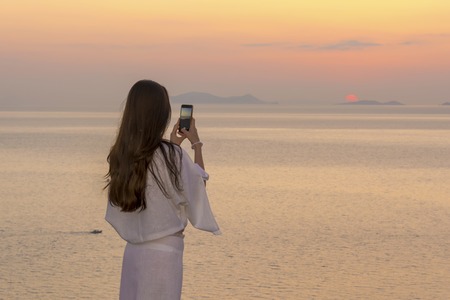 beautiful brunette taking pictures of the sunset in Santorini. Girl in transparent white dress makes the photograph.の写真素材