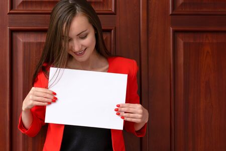 young smiling business woman in red suit with a white piece of paper on the background of wooden doors in the office. Place to test, copy space, mock upの写真素材