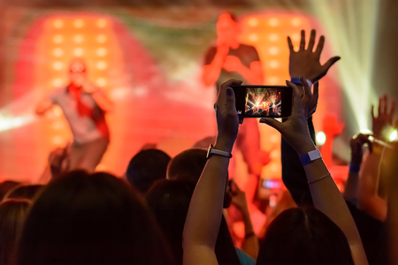silhouette of hands with a smartphone on the background of the singing artists in the light of the red lightsの写真素材
