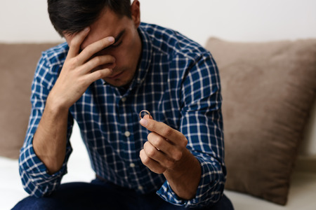 Close up of a sad husband after divorce, holding the wedding ring in a house interior. Selective...